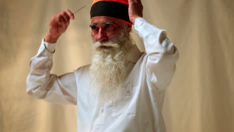 Fotografía-De-Estudio-De-Un-Hombre-Sikh-Mayor-Con-Barba-Que-Usa-Una-Aguja-De-Salai-Mientras-Se-Pone-Un-Turbante-Contra-Un-Fondo-Liso,-Filmada-En-Tiempo-Real